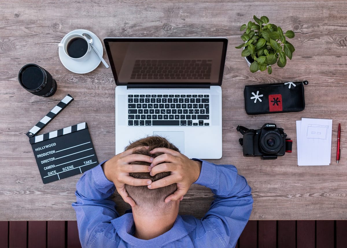 A top down image of a man clutching his head in front of his laptop, camera, notes, camera lens, plant, a cup of coffee, and a clapperboard.
