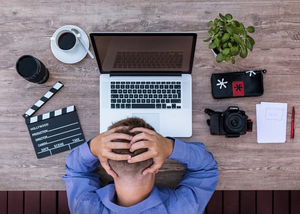 A top down image of a man clutching his head in front of his laptop, camera, notes, camera lens, plant, a cup of coffee, and a clapperboard.